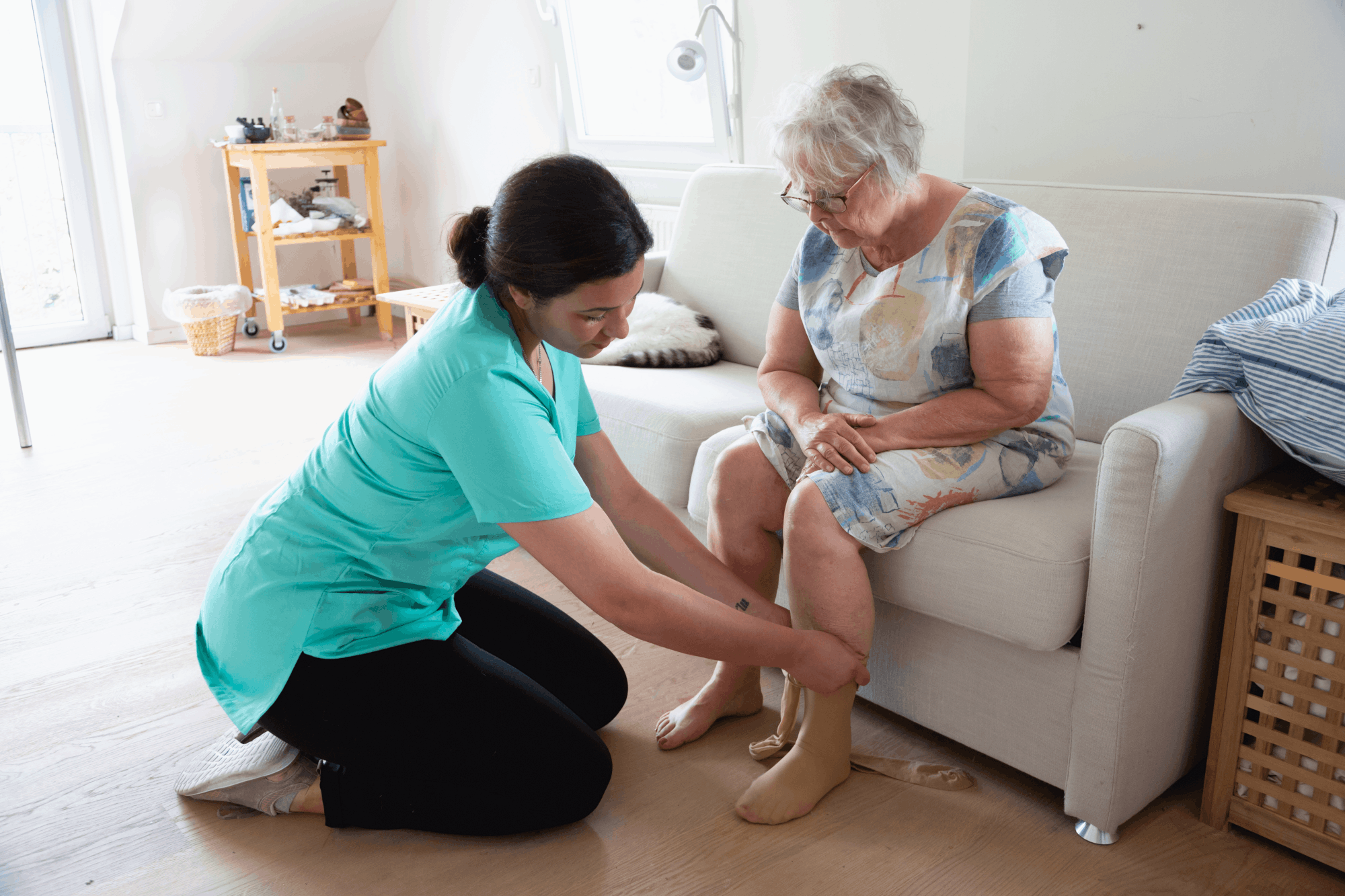 Pflegekraft hilft älterer Frau beim Anziehen von Kompressionsstrümpfen im Wohnzimmer.