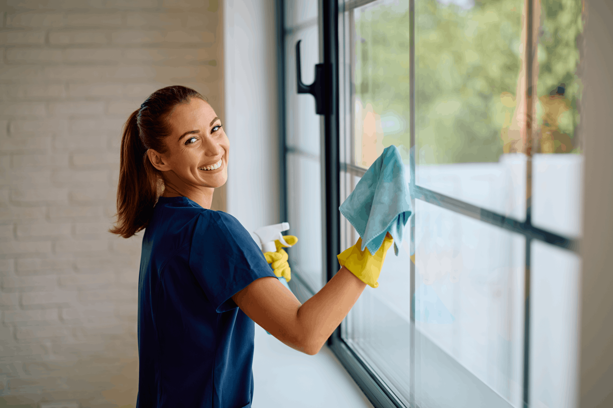 Frau reinigt ein Fenster mit Tuch und Spray und lächelt in die Kamera.